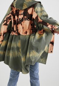 Giacca oversize tie-dye con toni della terra di verde, marrone e arancione, dotata di un cordino regolabile e numerose tasche.