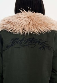 Giacca verde oliva con colletto in pelliccia sintetica beige e design di testo ricamato nero sulla schiena. Texture morbida e vestibilità ampia.