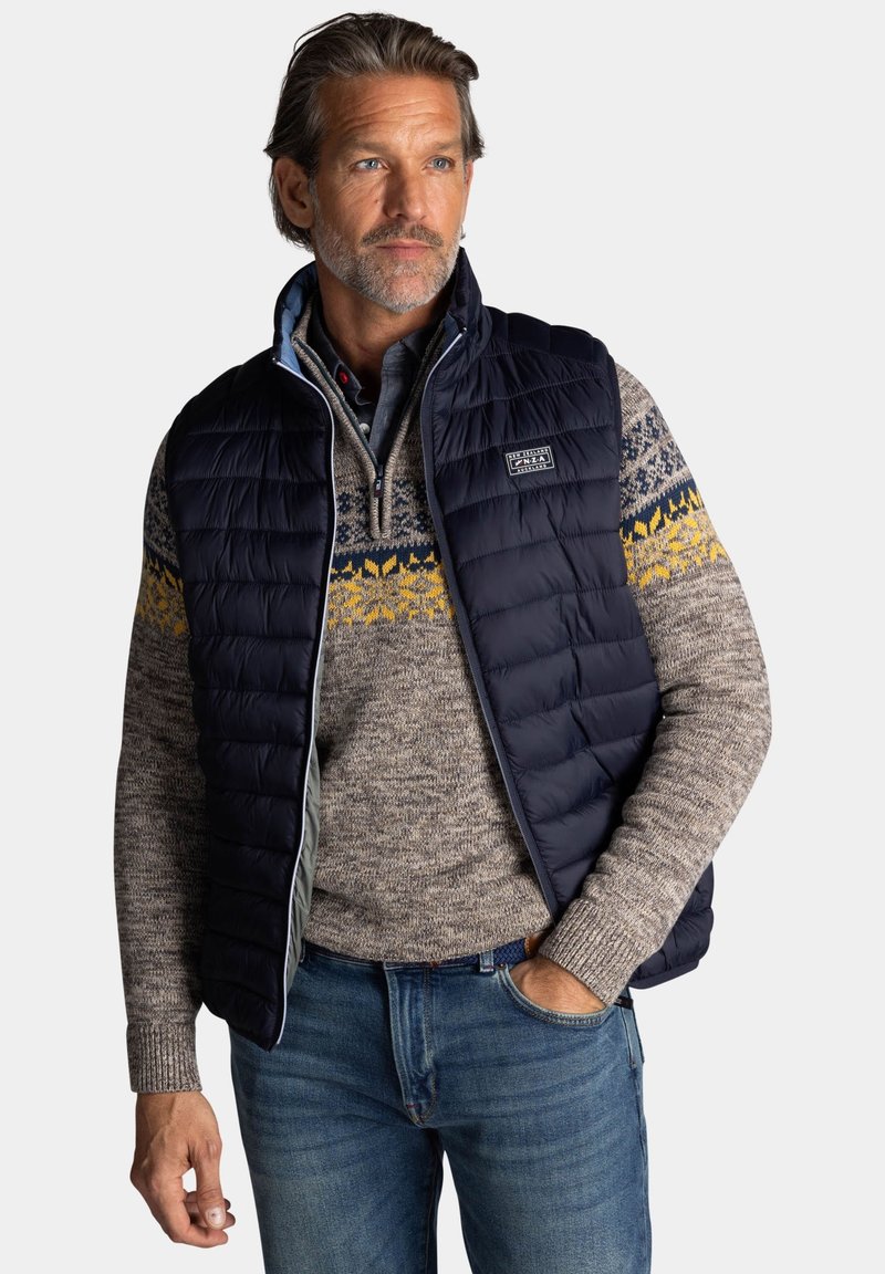 Marineblauwe puffer vest over een gemusterde bruine trui met gele en blauwe ontwerpen. De outfit is gecombineerd met een blauwe spijkerbroek.