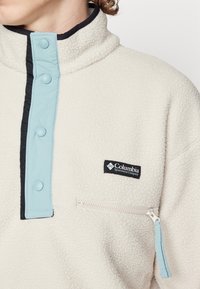 Pullover in pile crema con collo alto, dotato di una patta e bottoni blu chiaro, dettagli neri e una tasca con zip. Logo Columbia visibile.