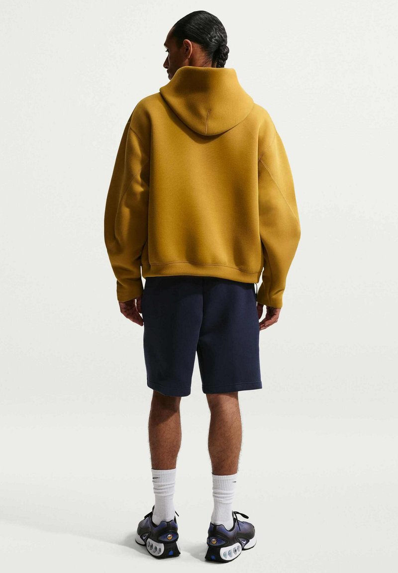 Persoon met donker haar in een lage paardenstaart draagt een mosterdkleurige hoodie, marineblauwe shorts, witte sokken en zwarte sneakers, met de rug naar de camera in een studio.