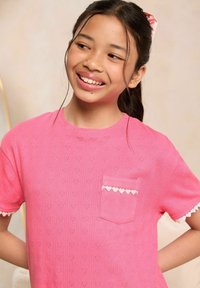 T-shirt rosa a maniche corte con motivi a forma di cuore, taschino frontale con bordo scallop e una finitura morbida e testurizzata.