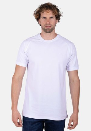 Hombre de pelo rizado de pie mirando al frente, lleva una camiseta blanca lisa de manga corta y vaqueros oscuros sobre un fondo claro.