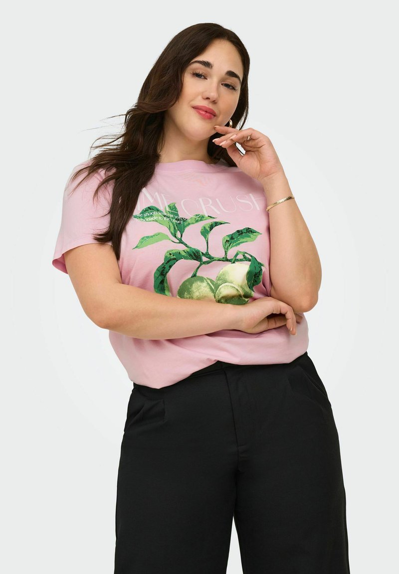 Femme aux longs cheveux foncés portant un t-shirt rose avec un imprimé de fruits verts et de feuilles et un pantalon noir, posant avec une main près de son visage.