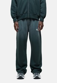 Donkergroene zip-up hoodie met voorzakken, gedragen met bijpassende, loszittende sweatpants met een vervaagd logo aan de linkerkant.