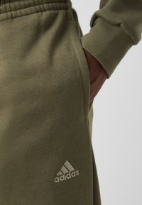 Pantaloni della tuta verde oliva realizzati in morbido tessuto, con cintura elastica, tasche laterali e un piccolo logo bianco Adidas sulla coscia.
