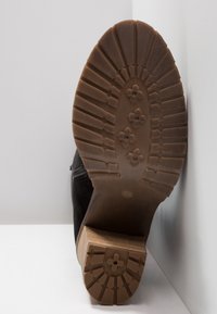Semelle d'une botte noire avec une épaisse semelle en caoutchouc, motifs floraux sur la semelle et talon en bois empilé vu de dessous sur fond blanc.
