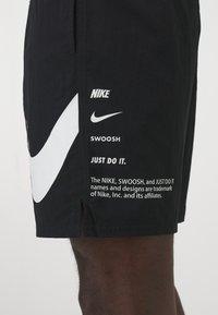 Mustat shortsit, joissa on suuri valkoinen Nike Swoosh -logo, lisäbrändäyselementtejä ja painatettu teksti vasemmassa lahkeessa. Kevyt kangas.