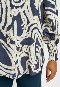 Blusa navy con motivo astratto bianco e azzurro chiaro, tessuto morbido, maniche a sbuffo e polsini arricciati.