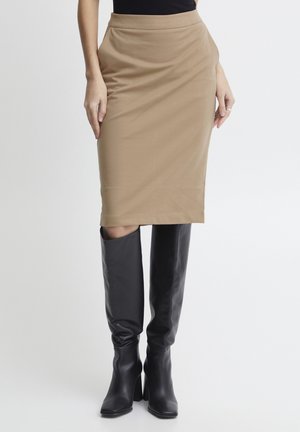 Femme portant une jupe crayon beige longueur genou et des bottes noires montantes à talons, debout devant un fond blanc uni.