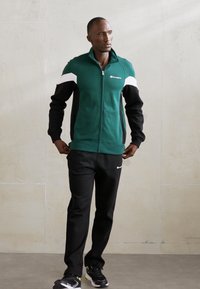 Giacca verde con zip, con accenti bianchi e neri, abbinata a pantaloni neri e sneakers nere con accenti neon. Tessuto morbido, design atletico.