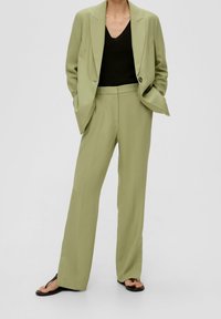 Costume ajusté vert clair avec un blazer structuré et un pantalon droit, associé à un top noir à col en V et des tongs noires.