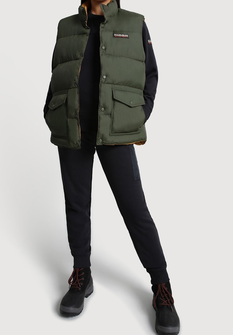 Personne portant une veste matelassée vert olive avec poches avant par-dessus un sweat-shirt noir et un pantalon noir, debout les mains dans les poches.