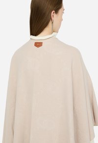 Poncho beige con una texture morbida, caratterizzato da un'etichetta in pelle marrone al colletto e un motivo circolare in rilievo su tutta la superficie.