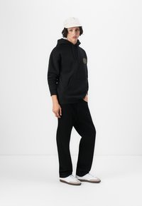 Black hoodie with pocket and graphic detail, paired with black pants and white sneakers. Model wears a light cap against a plain background.