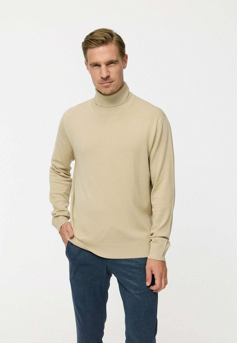 Bézs garbónyakú pulóver sima kötött anyagból, hosszú ujjakkal, bordás mandzsettákkal és szegéllyel, navy kék nadrággal viselve.