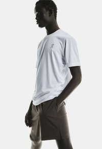 Mann in einem lockeren weißen Kurzarmhemd und braunen Shorts mit Händen in den Taschen, nach links auf einem hellen, einfarbigen Hintergrund gerichtet.