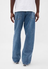 Modre kavbojke širokih hlačnic z izpranim videzom, z dvema zadnjima žepoma in surovim robom. Material izgleda debel in trpežen.