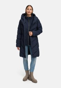 Jūras spēku pūkains mētelis ar kapuci, ar kapitālu dizainu un priekšējiem pogām. Apvienots ar gaišām džinsu biksēm un pelēkiem potīšu zābakiem.