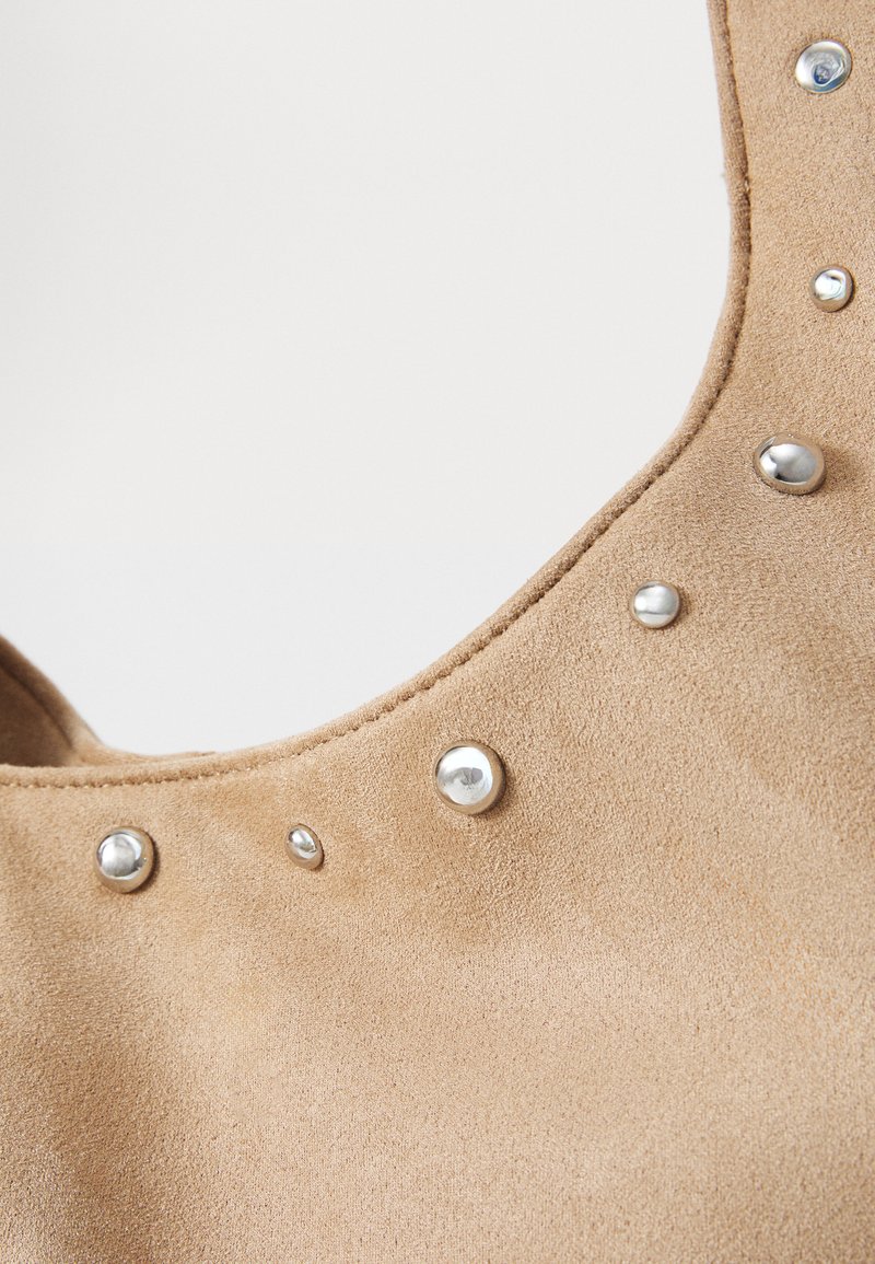 Bolso de ante marrón claro con textura suave, que presenta tachuelas metálicas a lo largo del borde. La forma es curveada y tiene una abertura amplia.