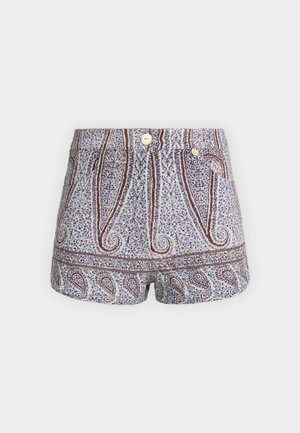 Mujer con camiseta blanca sin mangas, pantalones cortos de cintura alta con estampado, suéter negro atado alrededor de los hombros, calcetines blancos y zapatos planos negros, de pie sobre fondo blanco.