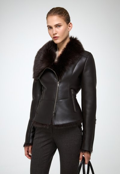 Donna che indossa una giacca di pelle nera con un ampio colletto e polsini di pelliccia, pantaloni scuri, tiene una borsa nera su uno sfondo semplice.