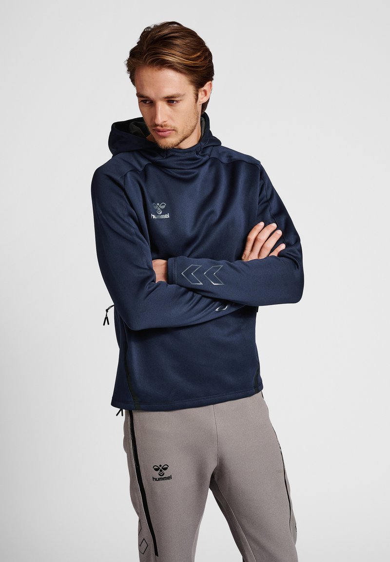 Mand med foldede arme iført navy hættetrøje og grå joggingbukser med hummel-logoer mod ensfarvet grå baggrund.