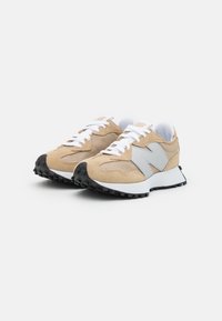 Pár béžových a bílých tenisek New Balance s černými texturovanými podrážkami, bílými tkaničkami a síťovinovými detaily vystavený na bílém pozadí.