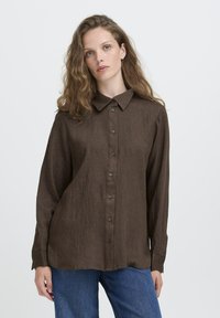Eine Frau mit welligem Haar, die ein braunes, durchgeknöpftes Langarmhemd und eine blaue Jeans trägt, vor einem schlichten weißen Hintergrund.