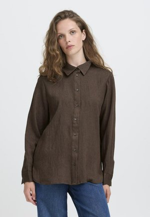 Femme avec des cheveux ondulés portant une chemise marron à boutons et des jeans bleus sur fond blanc.