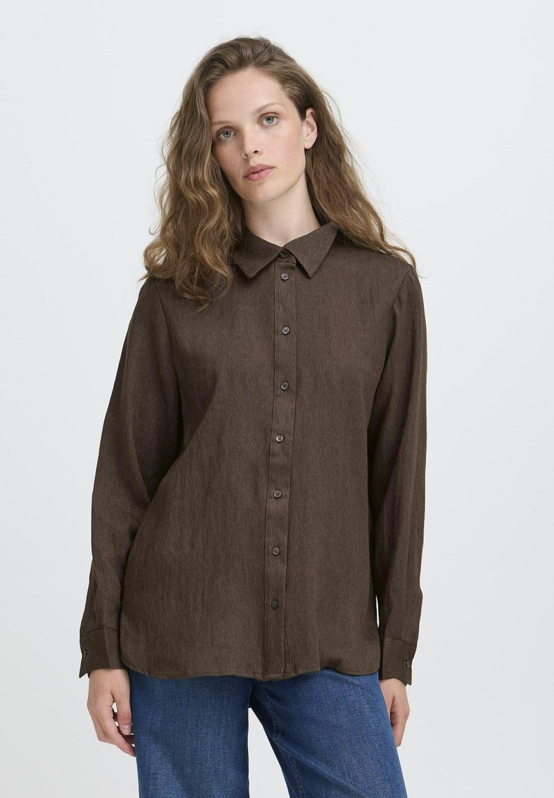 Eine Frau mit welligem Haar, die ein braunes, durchgeknöpftes Langarmhemd und eine blaue Jeans trägt, vor einem schlichten weißen Hintergrund.