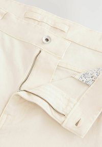 Beige cotton pants with a metal zipper and button closure. Features a patterned fabric lining visible at the zipper. Stitching detail along edges.