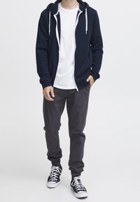 Sudadera con cremallera en color navy con cordones blancos, bolsillos frontales, llevada sobre una camiseta blanca, combinada con pantalones grises y zapatillas negras con cordones blancos.