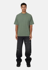 T-shirt oversize verde oliva in cotone, abbinato a jeans neri strappati con dettagli di schizzi di vernice. Sneakers nere completano il look.