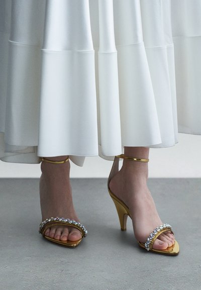 Sandali con tacco alto dorati con decorazioni gioiello, design a punta aperta e cinturino alla caviglia, indossati con una gonna bianca fluida.