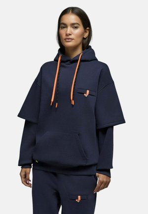 Femme portant un sweat à capuche bleu marine oversize et un pantalon assorti avec des cordons orange et petits détails de poche, debout devant un fond uni.