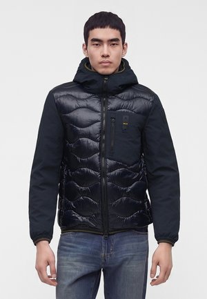 Giovane uomo che indossa una giacca trapuntata nera con cappuccio e zip frontale, abbinata a jeans blu, in piedi davanti a uno sfondo semplice.