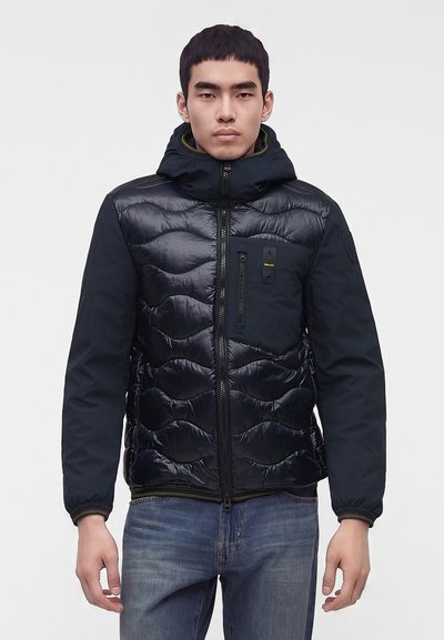 Jeune homme portant une veste matelassée noire avec une capuche et une fermeture éclair à l'avant, associée à un jean bleu, debout devant un fond uni.