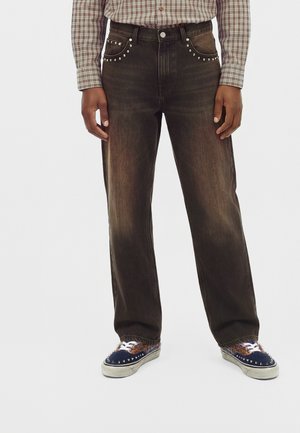 Homme portant un jean noir délavé avec des clous argentés sur les poches avant, associé à des baskets bleues et marron à clous et une chemise marron à carreaux.