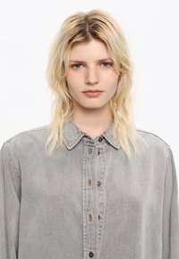 Graues Denim-Hemd mit spitzem Kragen, Knopfverschluss und braunen Knöpfen. Es hat lange Ärmel und eine lockere Passform. Strukturierter Stoff.