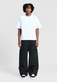 Uomo con capelli ricci che indossa una maglietta bianca oversize, pantaloni cargo neri con più tasche e sneakers bianche e nere, in piedi dritto.