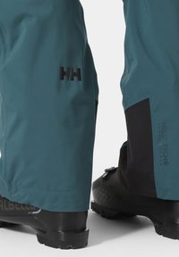 Vodootporne hlače u plavoj-zelenoj boji s crnim detaljima, sa logotipom i Helly Tech oznakom. Upotrebljeni materijal je gladak bez istaknutih tekstura.