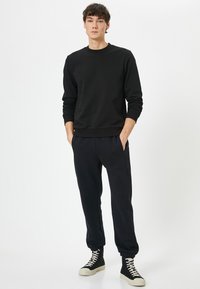 Sort sweatshirt med rund hals og ribbede manchetter, parret med mørke joggers. Footwear inkluderer høje sorte sneakers med hvide såler.