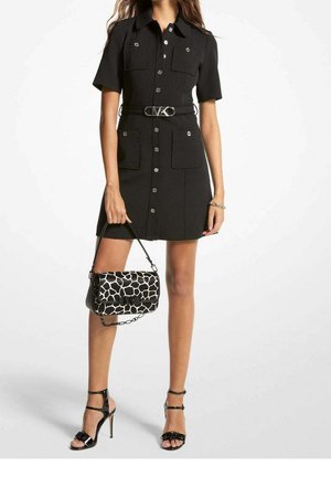 Femme portant une mini-robe noire boutonnée à manches courtes avec ceinture, tenant un sac à main noir et blanc à motifs, et portant des sandales noires à talons hauts.