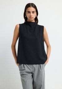 Femme aux cheveux bruns portant des boucles d'oreilles en argent, un haut sans manches noir et un pantalon plissé gris, se tenant les mains dans les poches.
