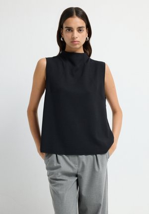 Donna con capelli scuri che indossa orecchini a cerchio argentati, una canotta nera senza maniche e pantaloni grigi a pieghe, in piedi con le mani in tasca.