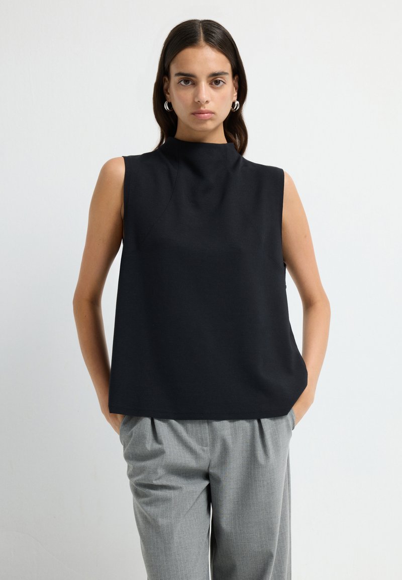 Femme aux cheveux bruns portant des boucles d'oreilles en argent, un haut sans manches noir et un pantalon plissé gris, se tenant les mains dans les poches.