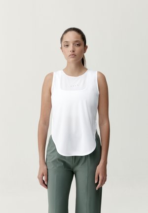 Junge Frau, die steht, trägt ein ärmelloses weißes Oberteil und hoch taillierte grüne Hosen vor einem schlichten hellgrauen Hintergrund.