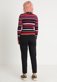 Persona con capelli rosa corti, girata di spalle, che indossa una maglietta a maniche lunghe a righe multicolori, jeans neri e sneakers verdi.
