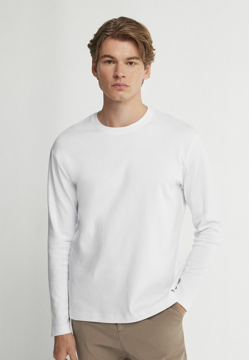 Weißes Langarm-T-Shirt aus einem weichen Baumwollmix, mit Rundhalsausschnitt und lockerer Passform, getragen mit hellen Hosen.
